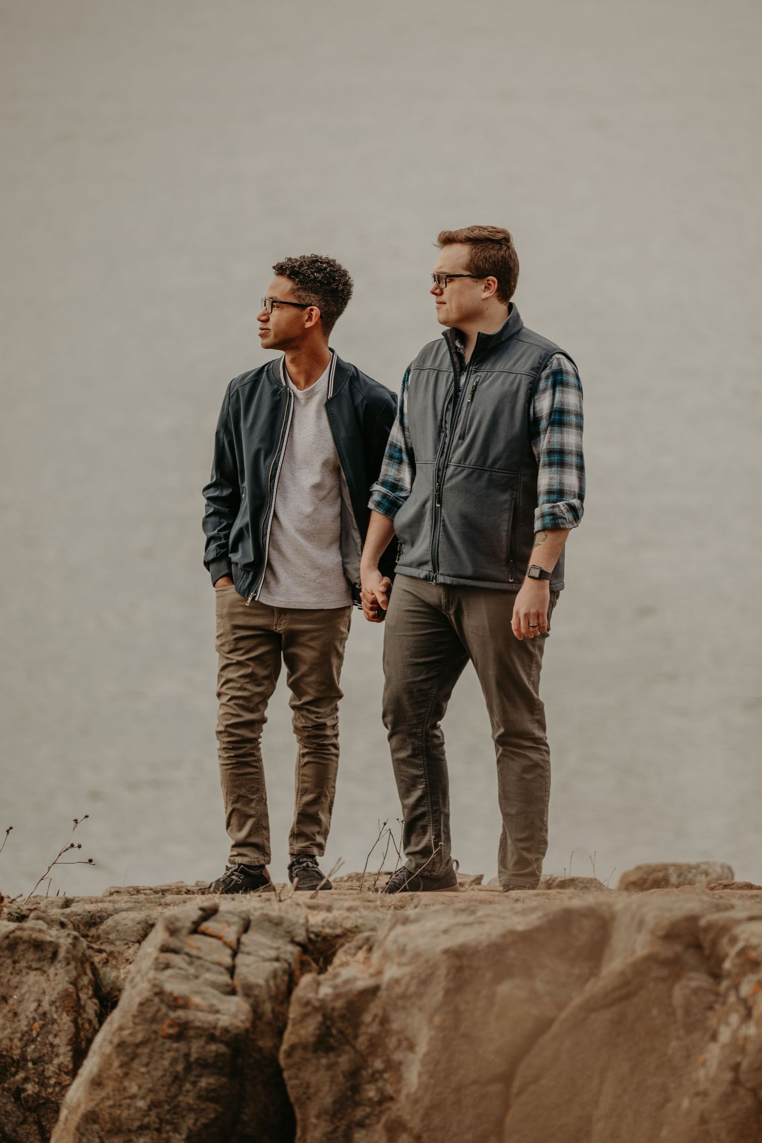 Two people in casual fall attire stand together on a rocky cliff edge against an overcast sky.