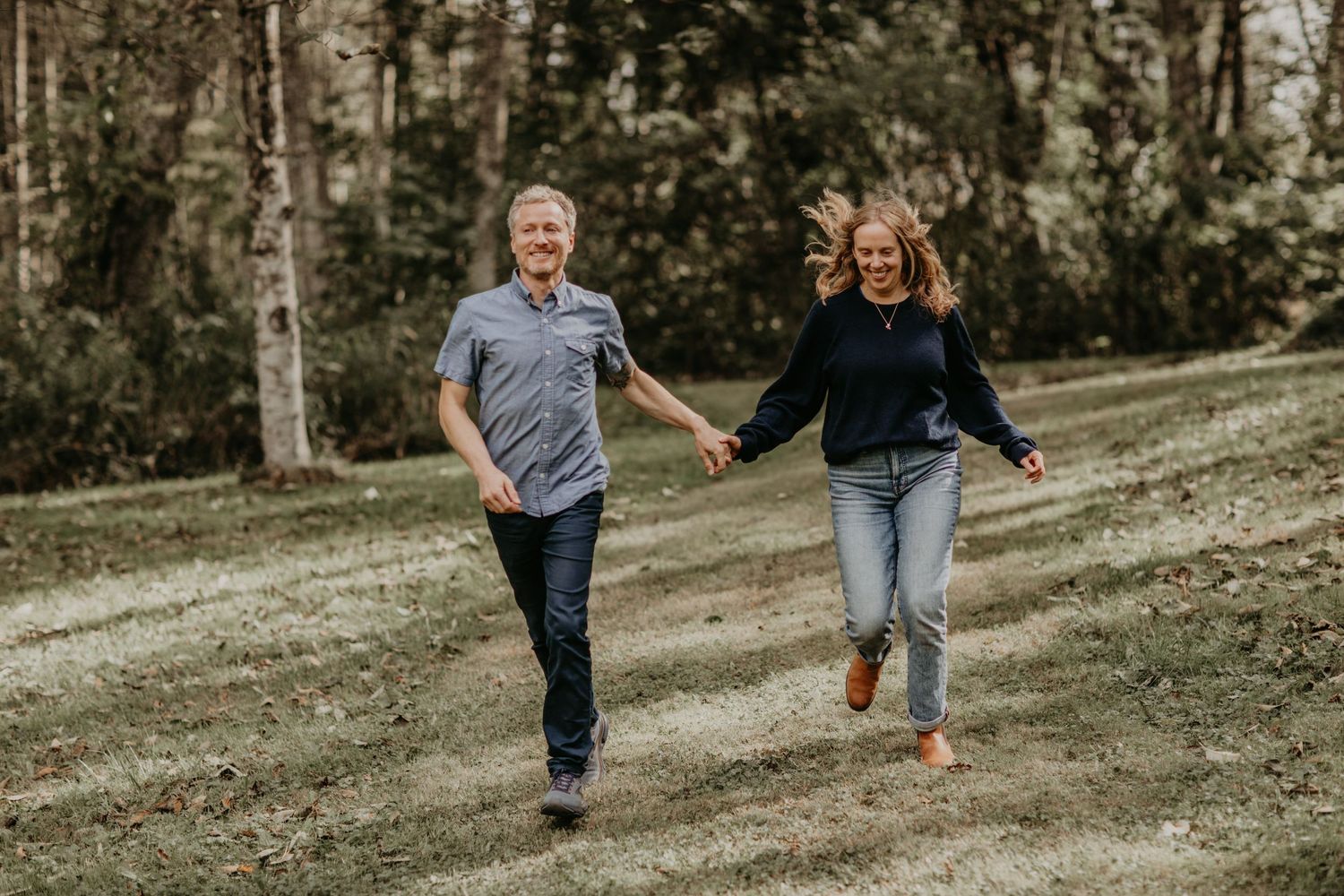Two people run playfully together down a grassy slope with trees in the background.