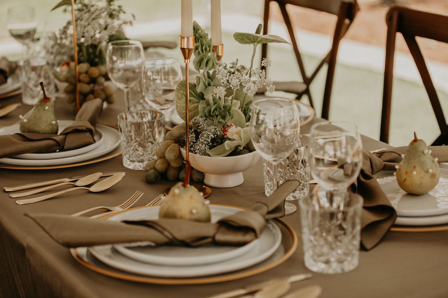 Rustic wedding table setting with beige linens, crystal glasses, white plates and natural greenery centerpieces.