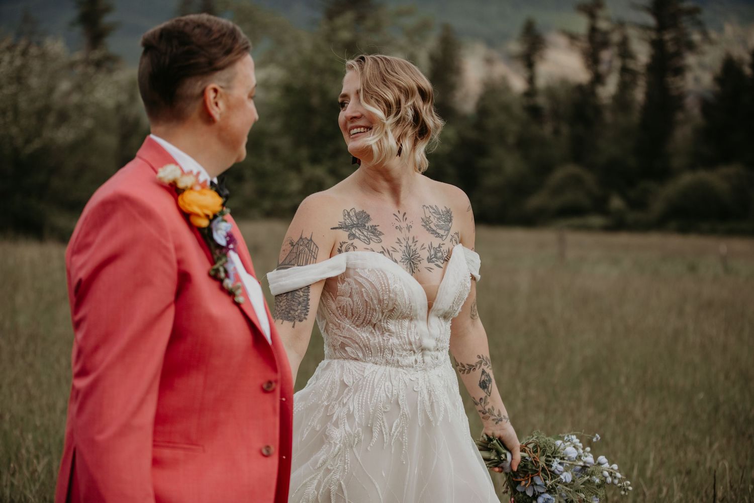 A couple in wedding attire stands in a rustic field with pine trees, the groom wearing a vibrant coral suit.