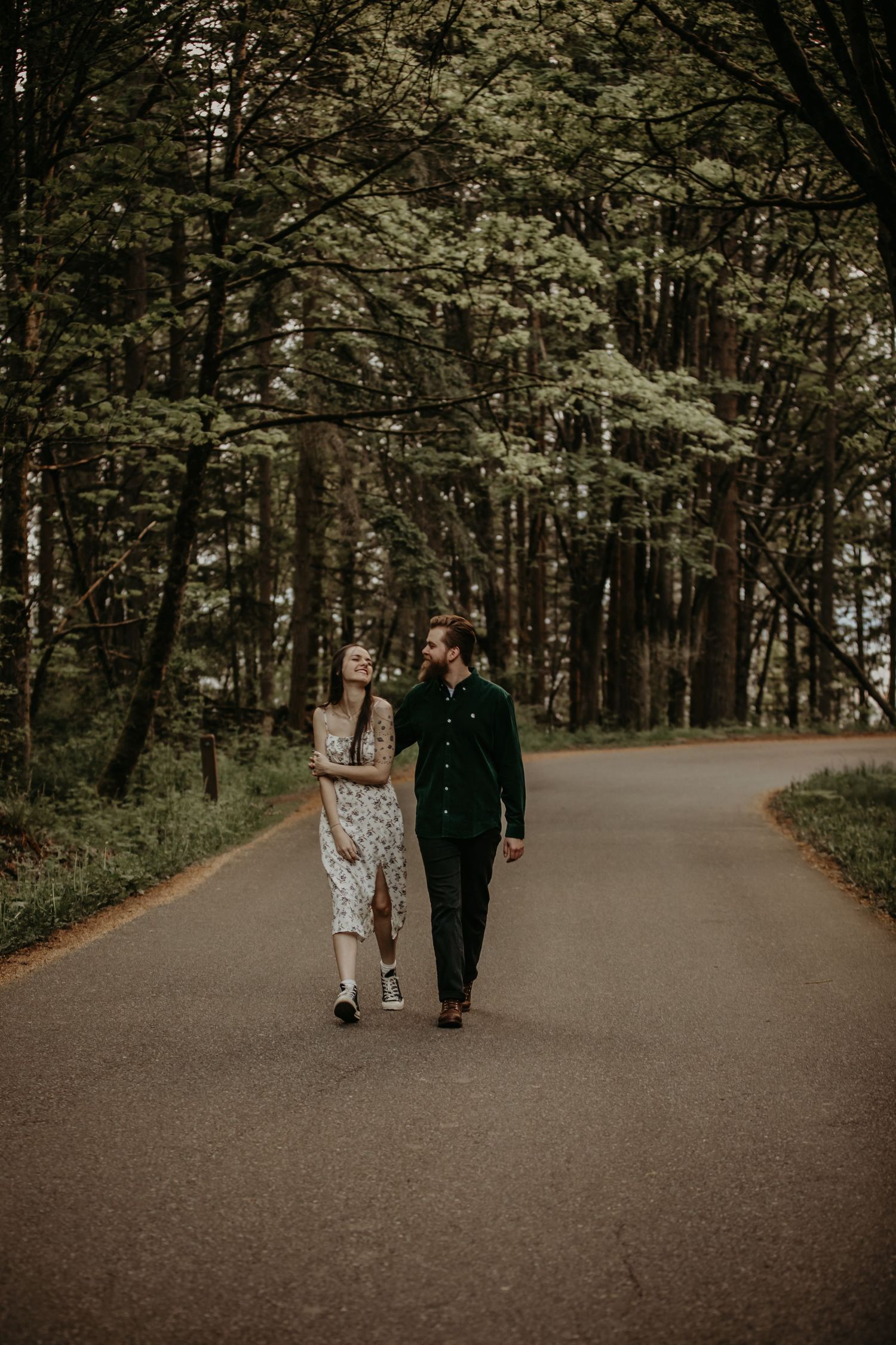 A couple walks together on a moody forest path with tall trees creating a romantic woodland atmosphere.