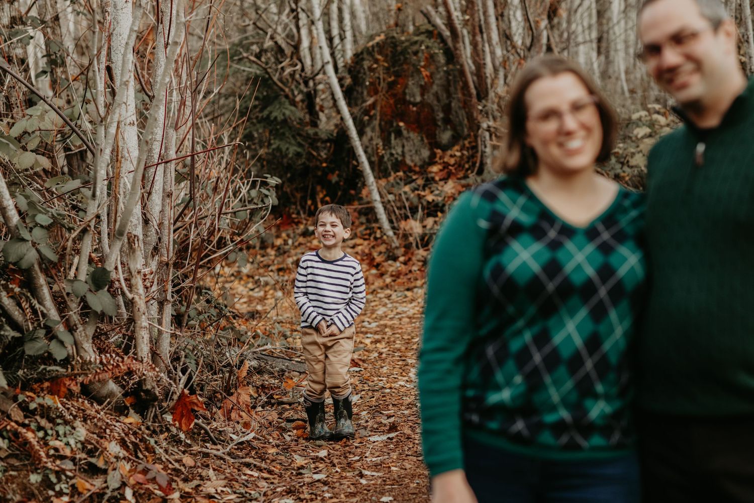 A child in striped clothing walks on a forest path while a couple stands blurred in the background.