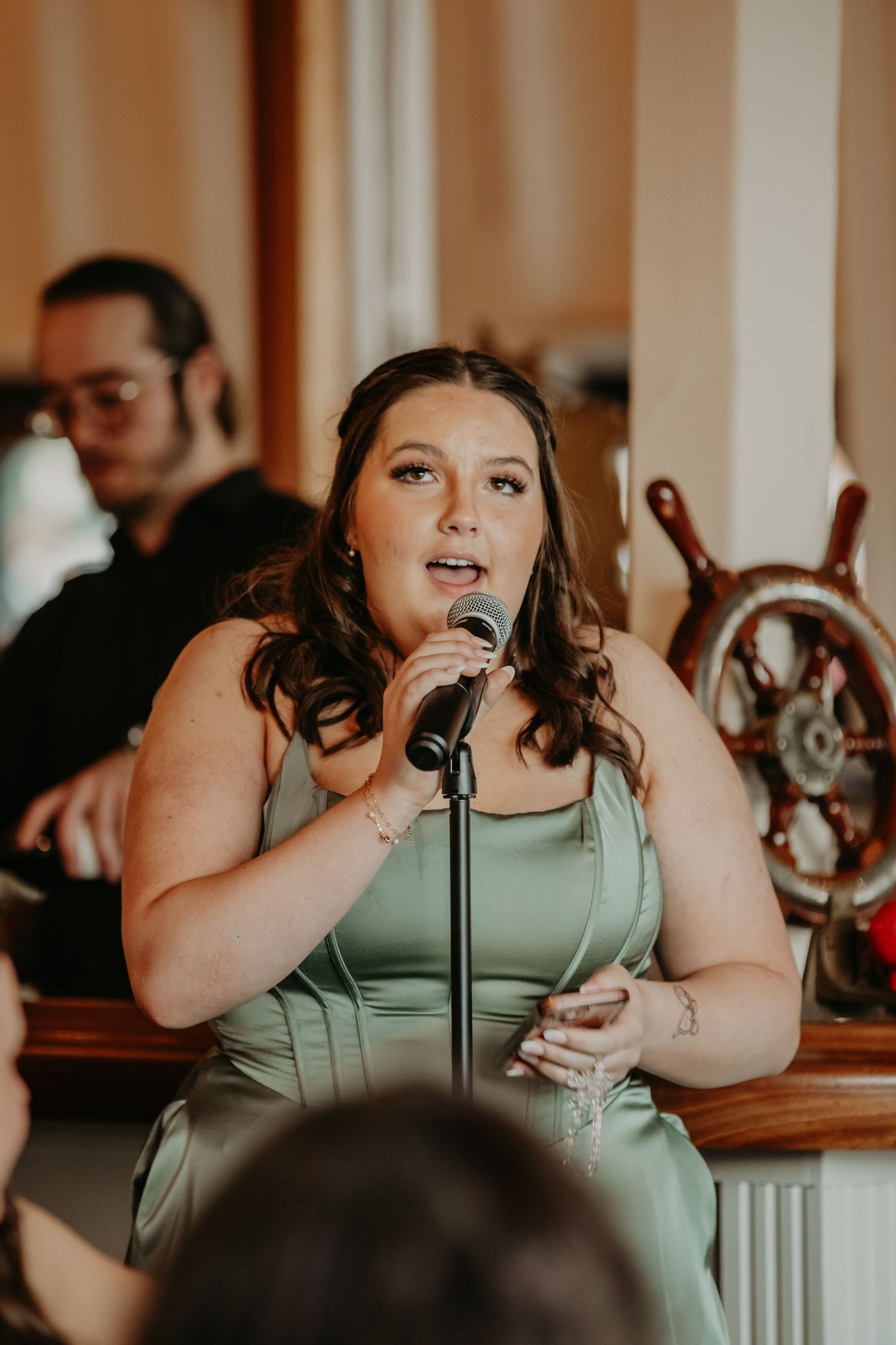 Someone in a sage green dress gives an emotional speech at an indoor event while holding a microphone and glass.
