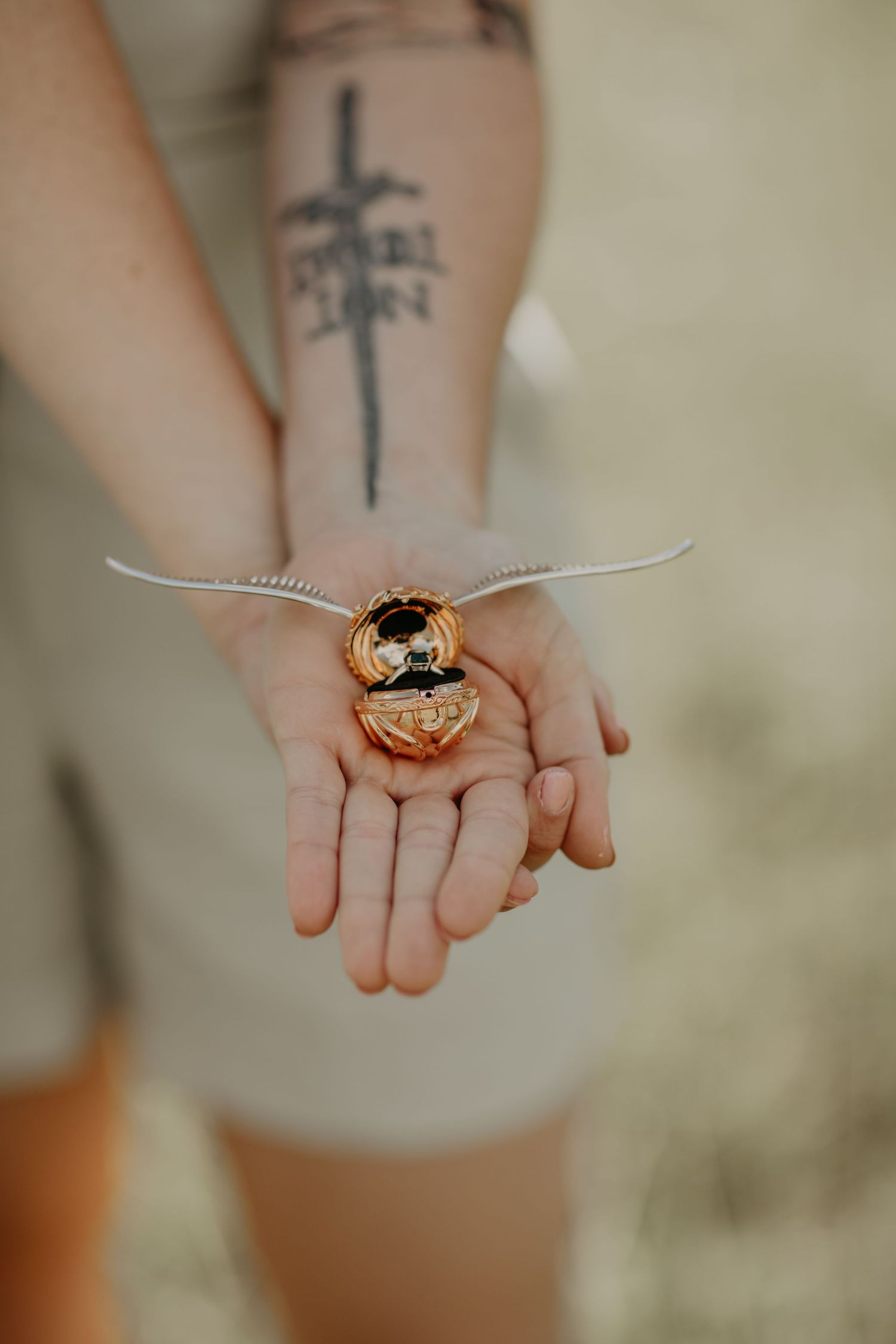 Golden snitch prop from Harry Potter held in an outstretched hand with a visible cross tattoo on the arm.