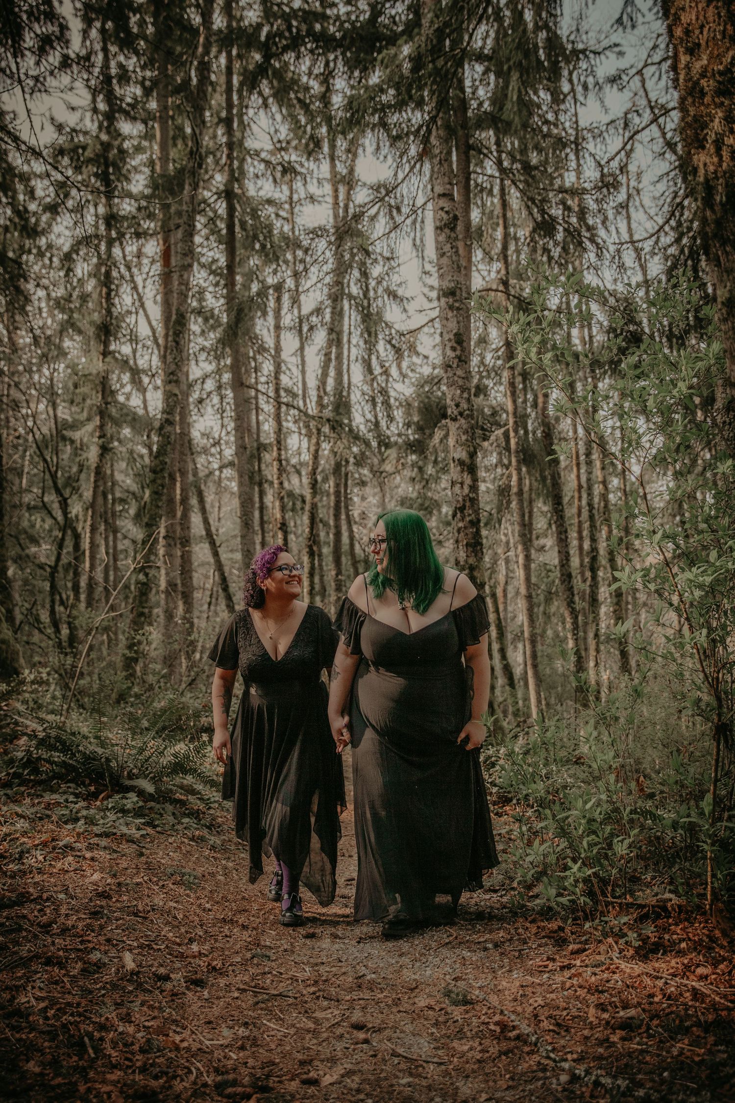 Two people wearing dark dresses walk together on a forest path surrounded by tall evergreen trees in moody lighting.