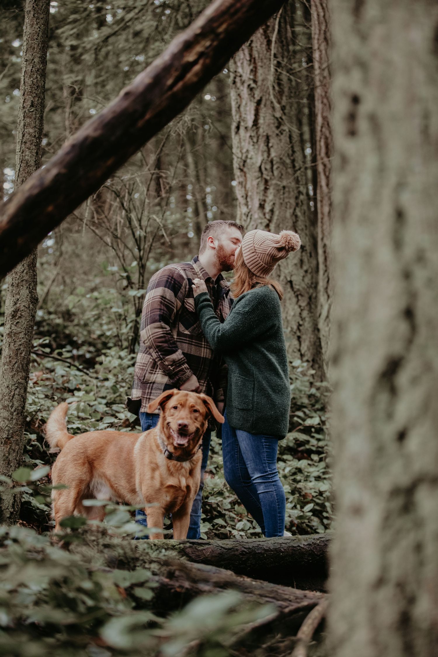 A couple shares an intimate moment in the woods with their golden retriever standing beside them.