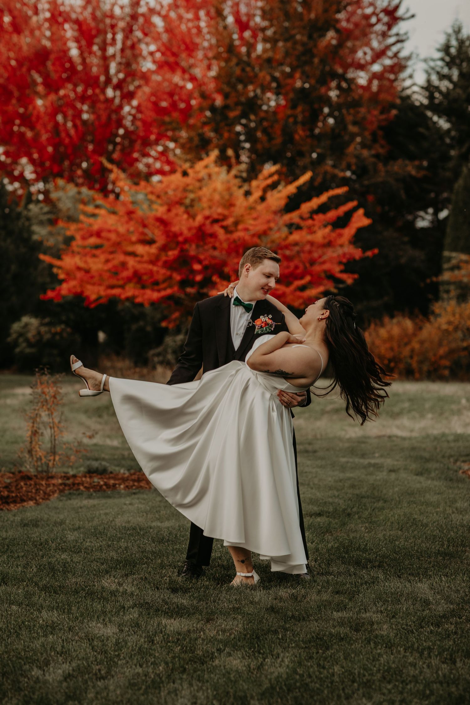 A romantic couple in wedding attire dances in front of vibrant red autumn foliage on a grassy lawn.