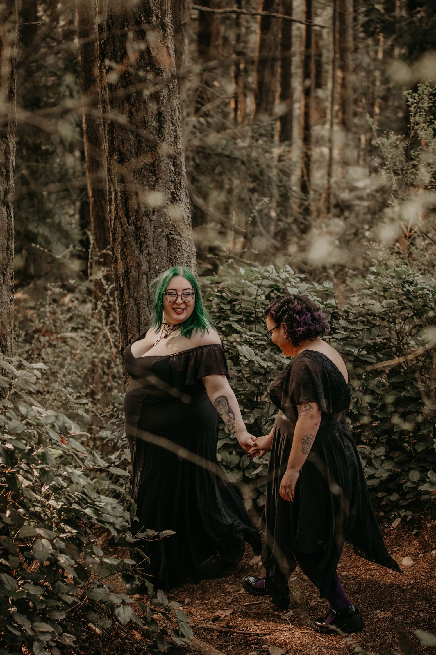 Two people in black dresses stand holding hands on a wooded forest path in moody natural lighting.