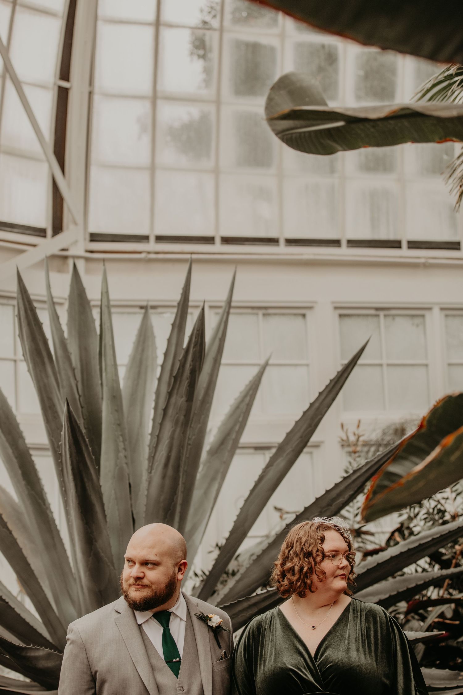 A moody, vintage-style photo showing a couple standing in front of large agave plants at a greenhouse or conservatory.