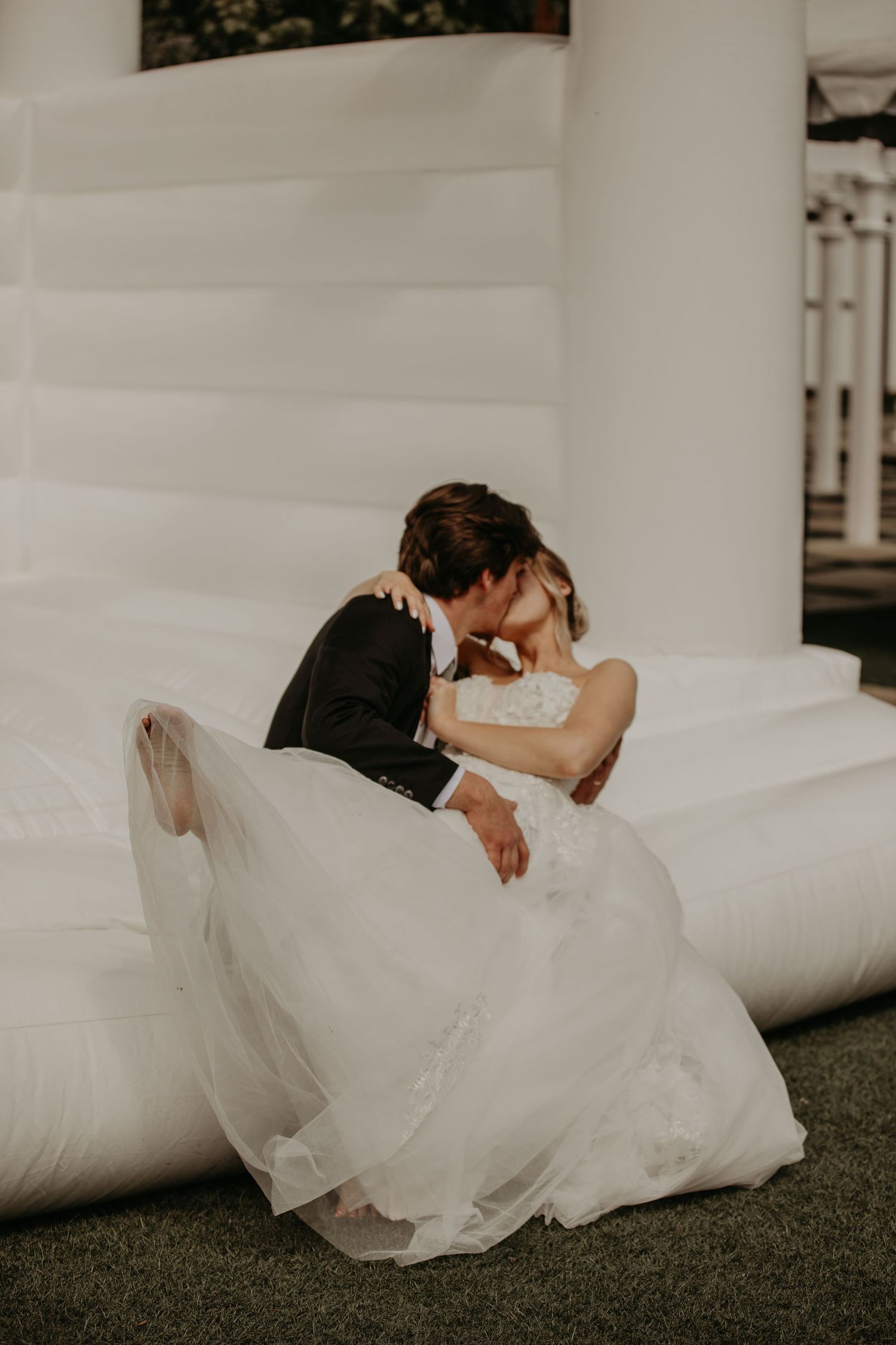A romantic wedding couple shares an intimate moment sitting together on white cushions outdoors.