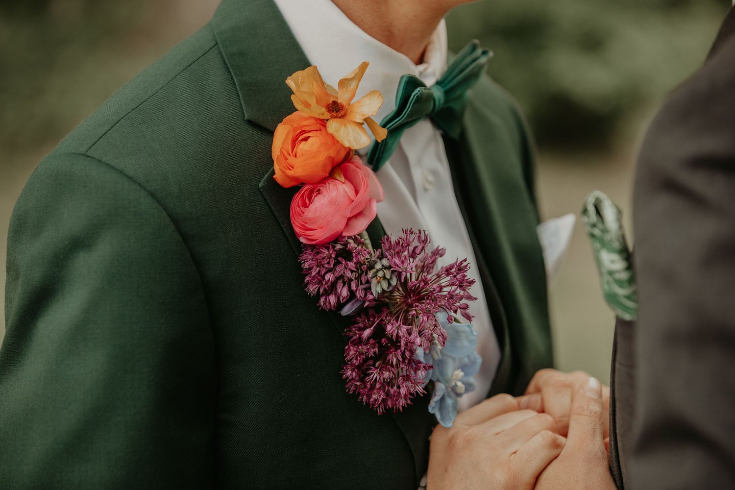A colorful boutonniere with orange and pink flowers pinned to a dark green suit jacket lapel.