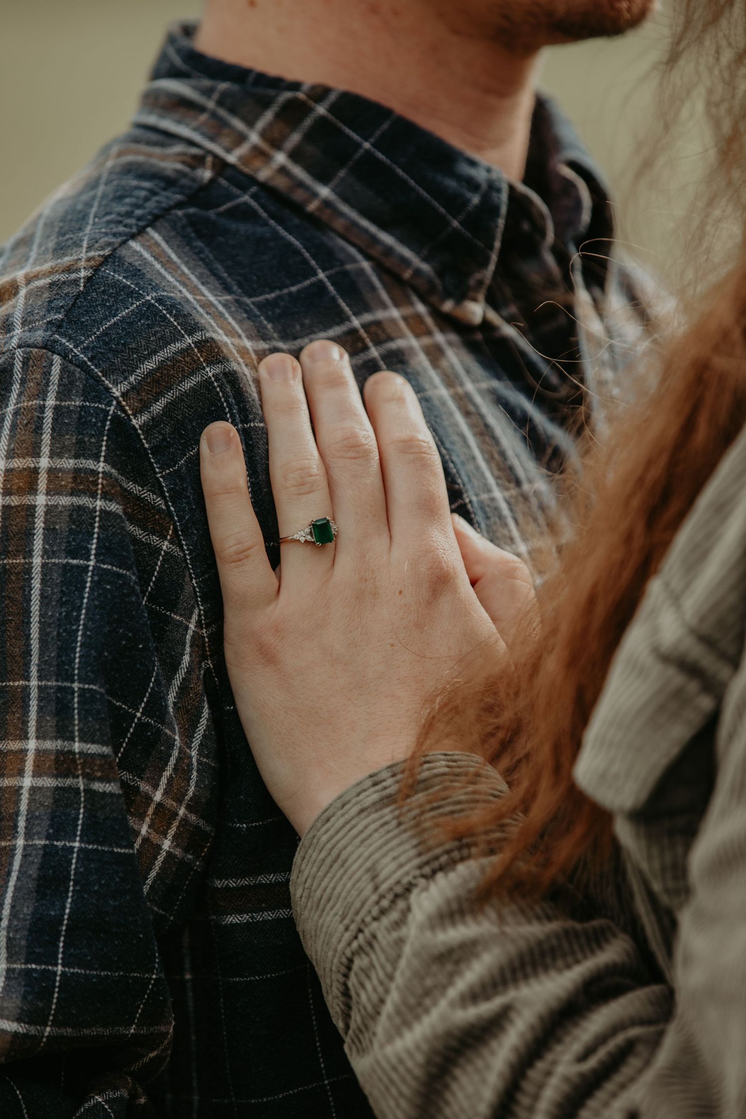 Close-up of a hand wearing an engagement ring resting on someone's plaid shirt covered shoulder.