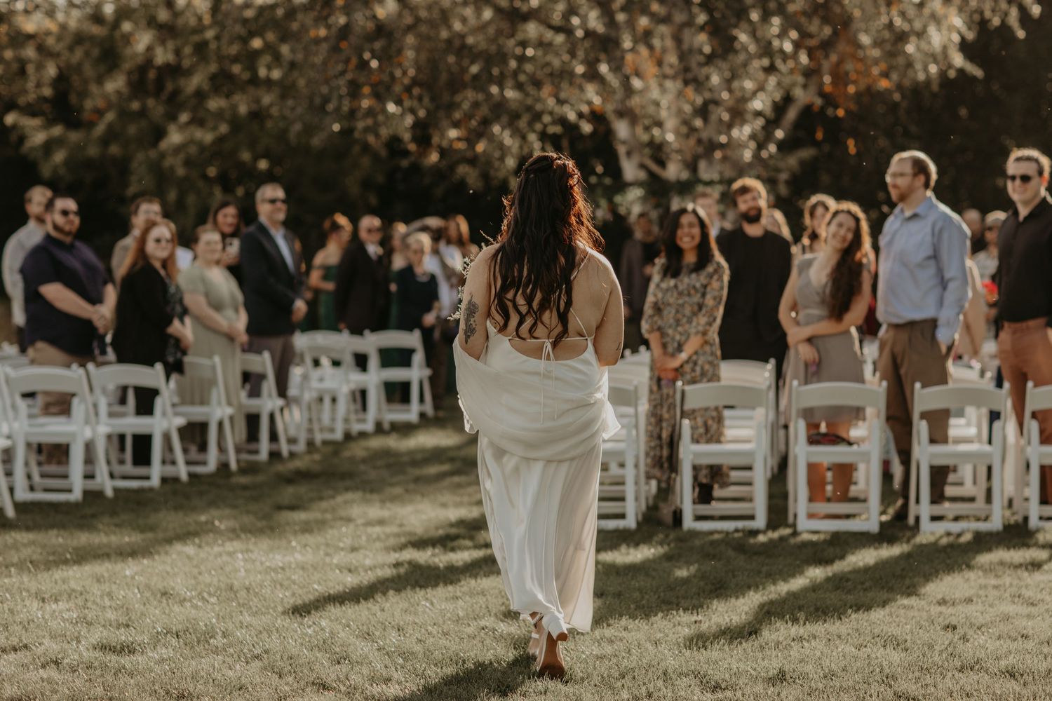 A backlit bride in a flowing white dress walks down an outdoor aisle lined with white chairs and guests at sunset.