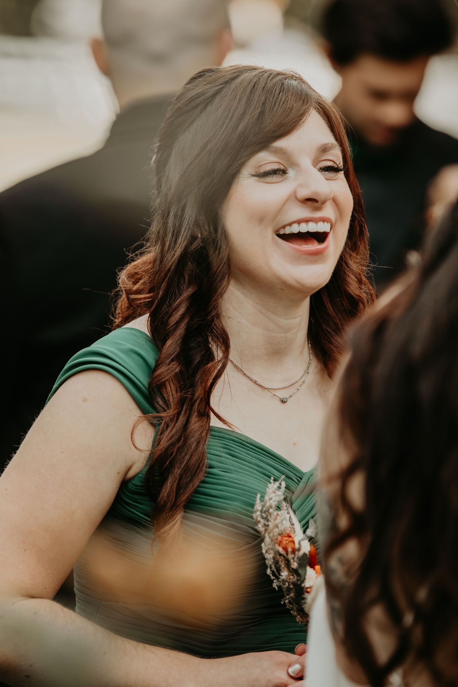Someone in an emerald green dress laughs joyfully at a wedding celebration, wearing a floral corsage.