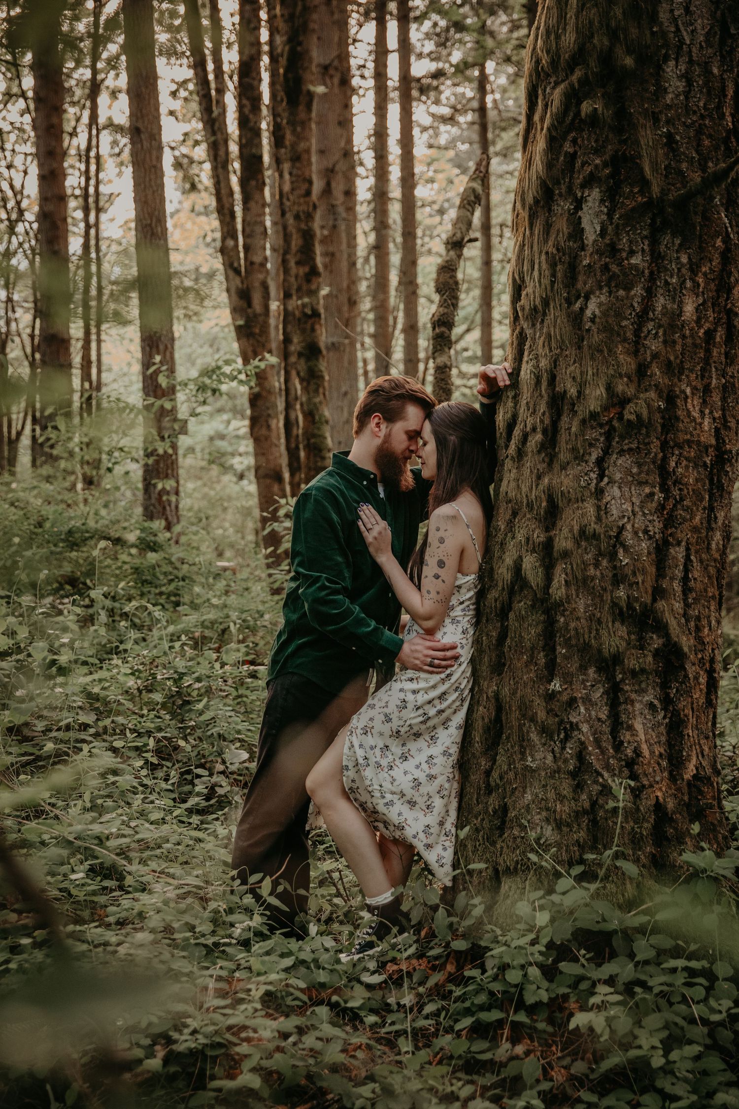 A romantic forest scene between two figures leaning against a large tree trunk in moody natural lighting.