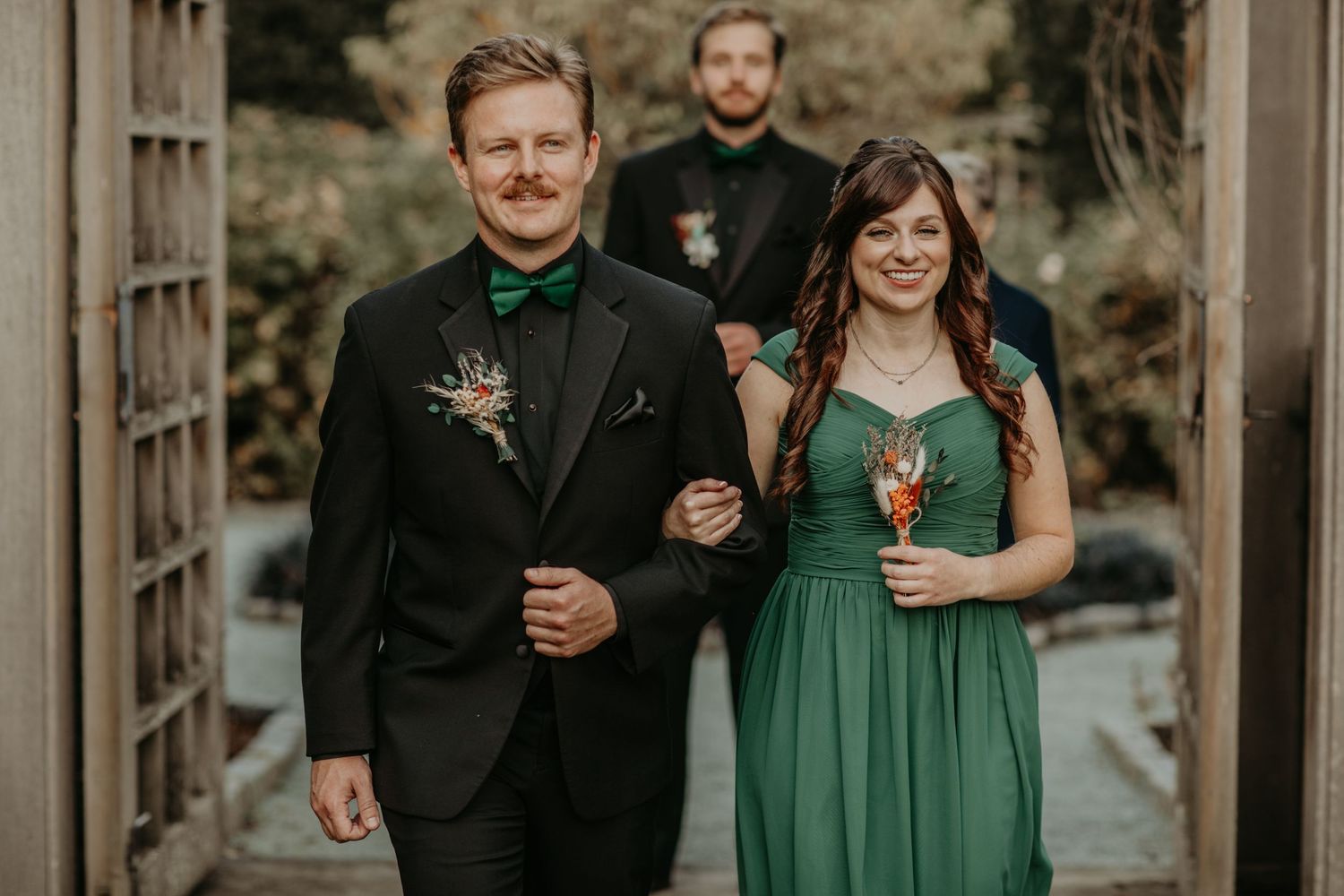 Wedding party members in formal attire walk through a garden arbor with blurred figure in background.