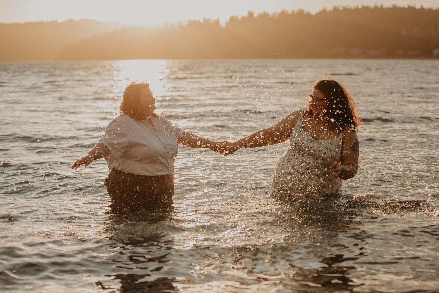 Two people splashing and holding hands in water at sunset while creating joyful moments together.