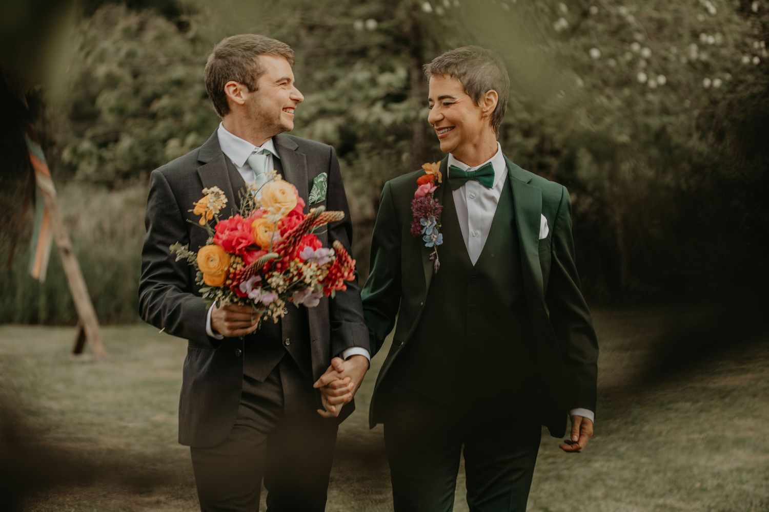 Two grooms in black tuxedos holding hands and laughing while one carries a vibrant coral bouquet in an outdoor setting.
