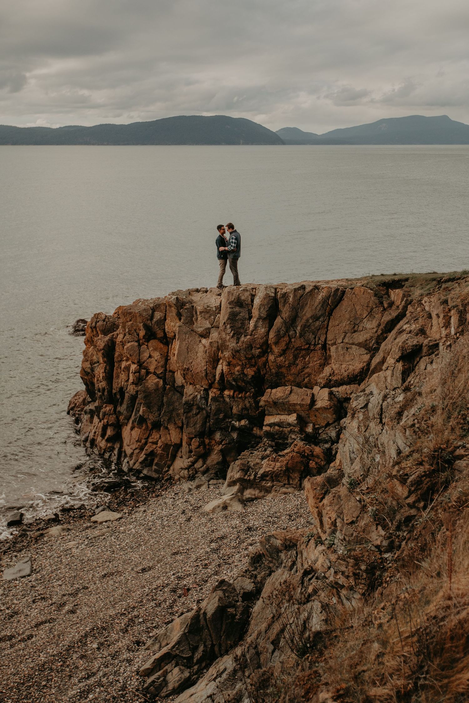 Couple embracing on dramatic coastal cliff edge overlooking misty mountains and gray waters on overcast day.