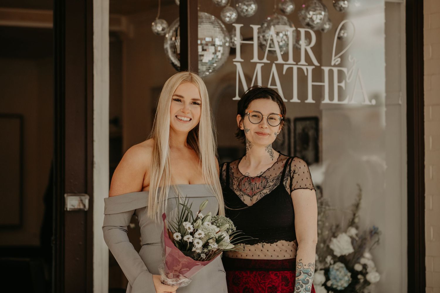 Two people standing in front of a hair salon window with a bridal bouquet and elegant formalwear.