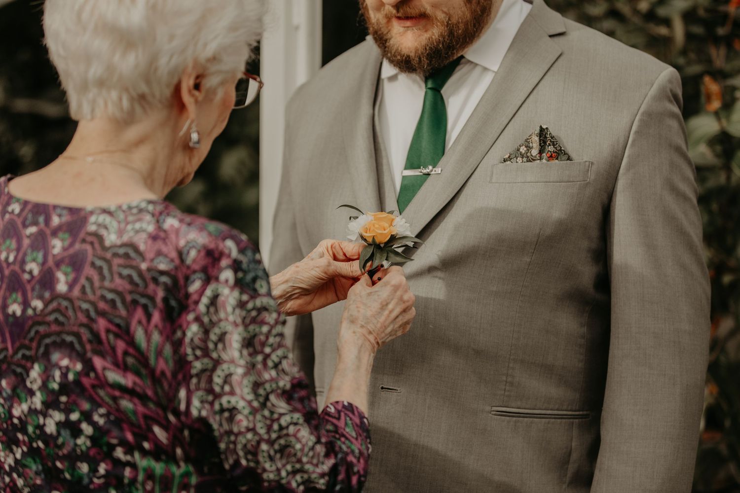 Person wearing a grey suit and green tie receives a boutonniere pin from someone in a floral print top.