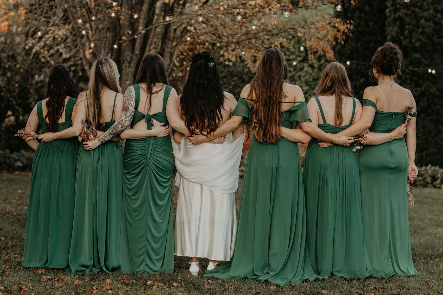 Bridesmaids wearing matching emerald green dresses stand with their arms around each other, facing away from the camera.