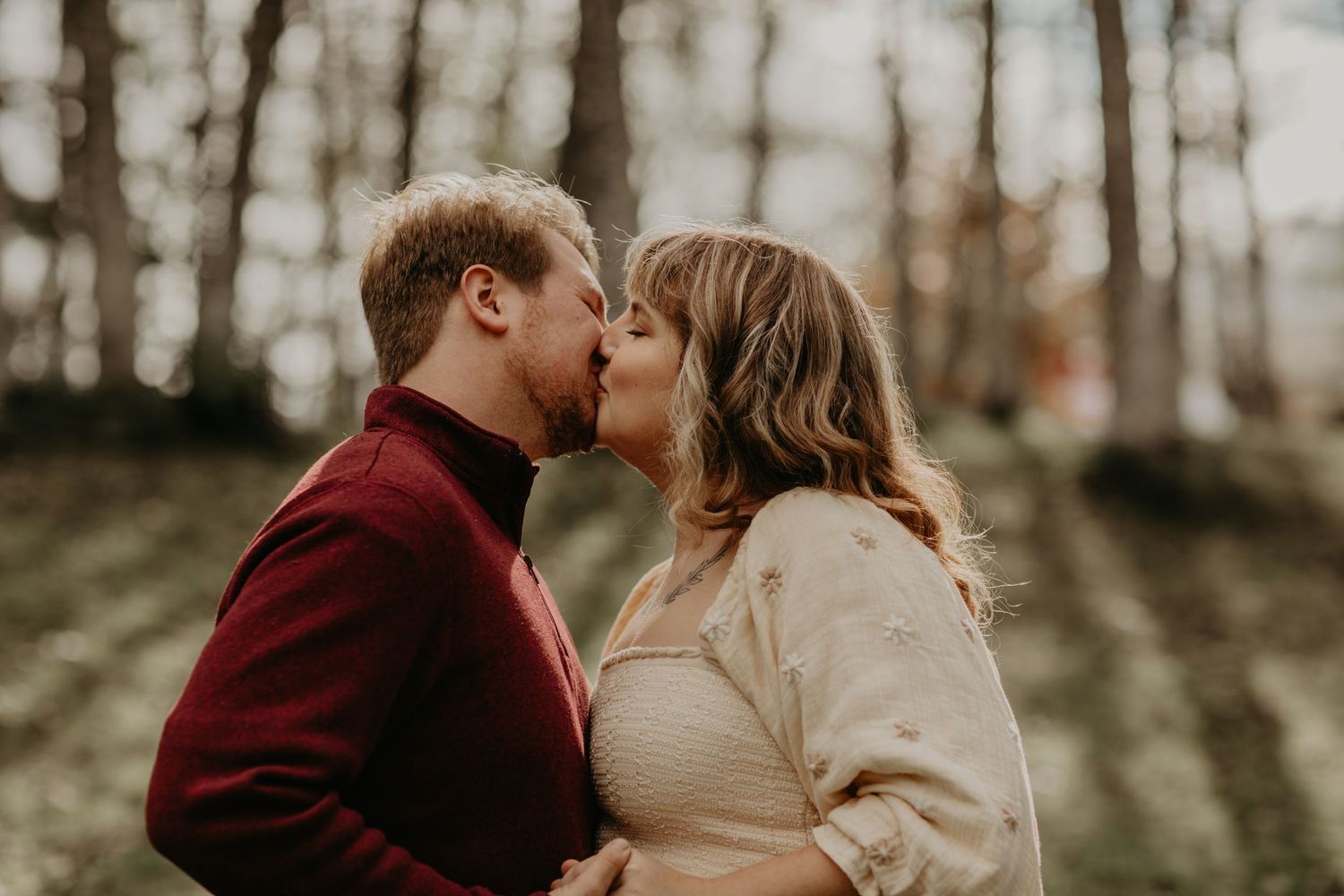A romantic autumn engagement photo with a couple sharing a kiss in a forest wearing burgundy and cream colored sweaters.
