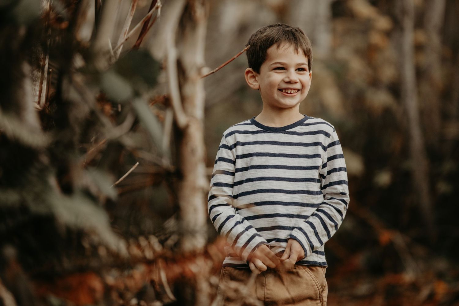A child in a striped sweater stands smiling outdoors in an autumn forest setting with blurred background.
