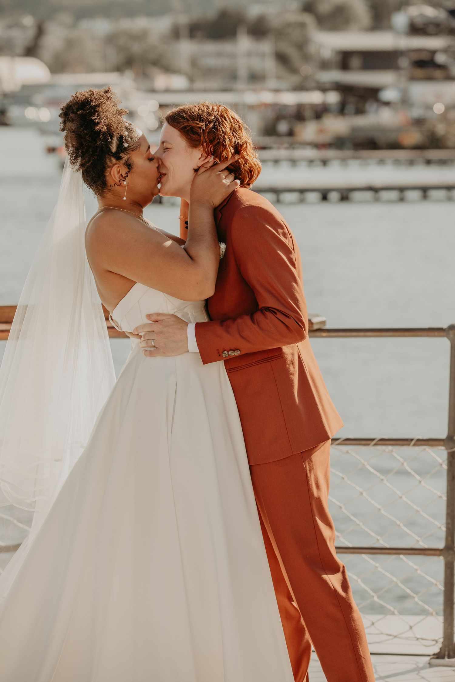 A romantic waterfront wedding kiss between a couple, one in white dress and one in rust-colored suit.