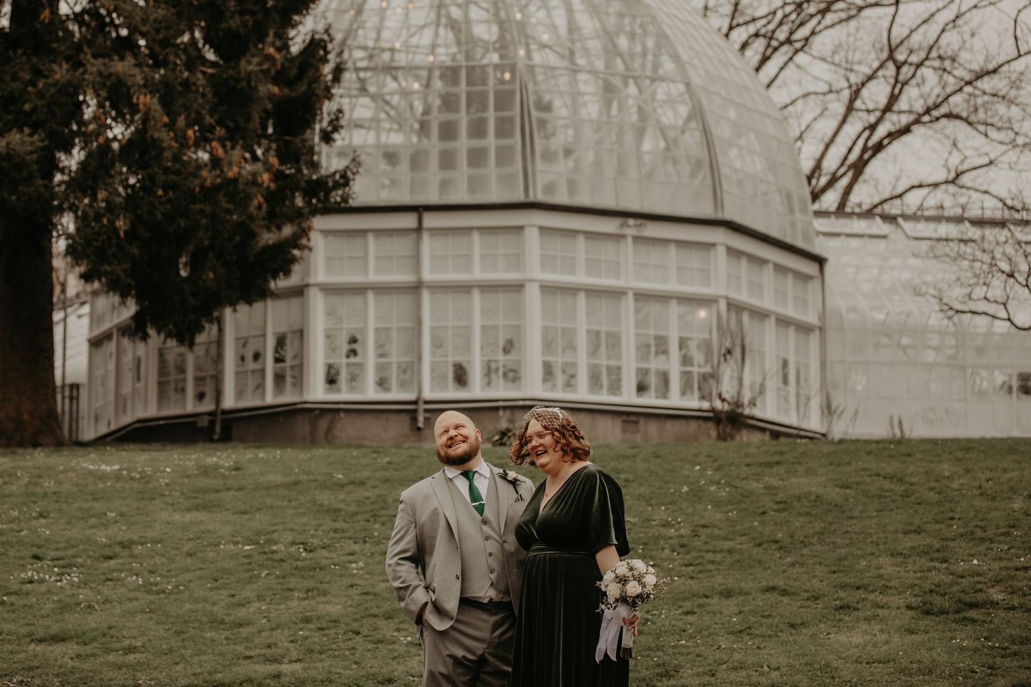 Wedding couple posed together in front of historic glass conservatory building.
