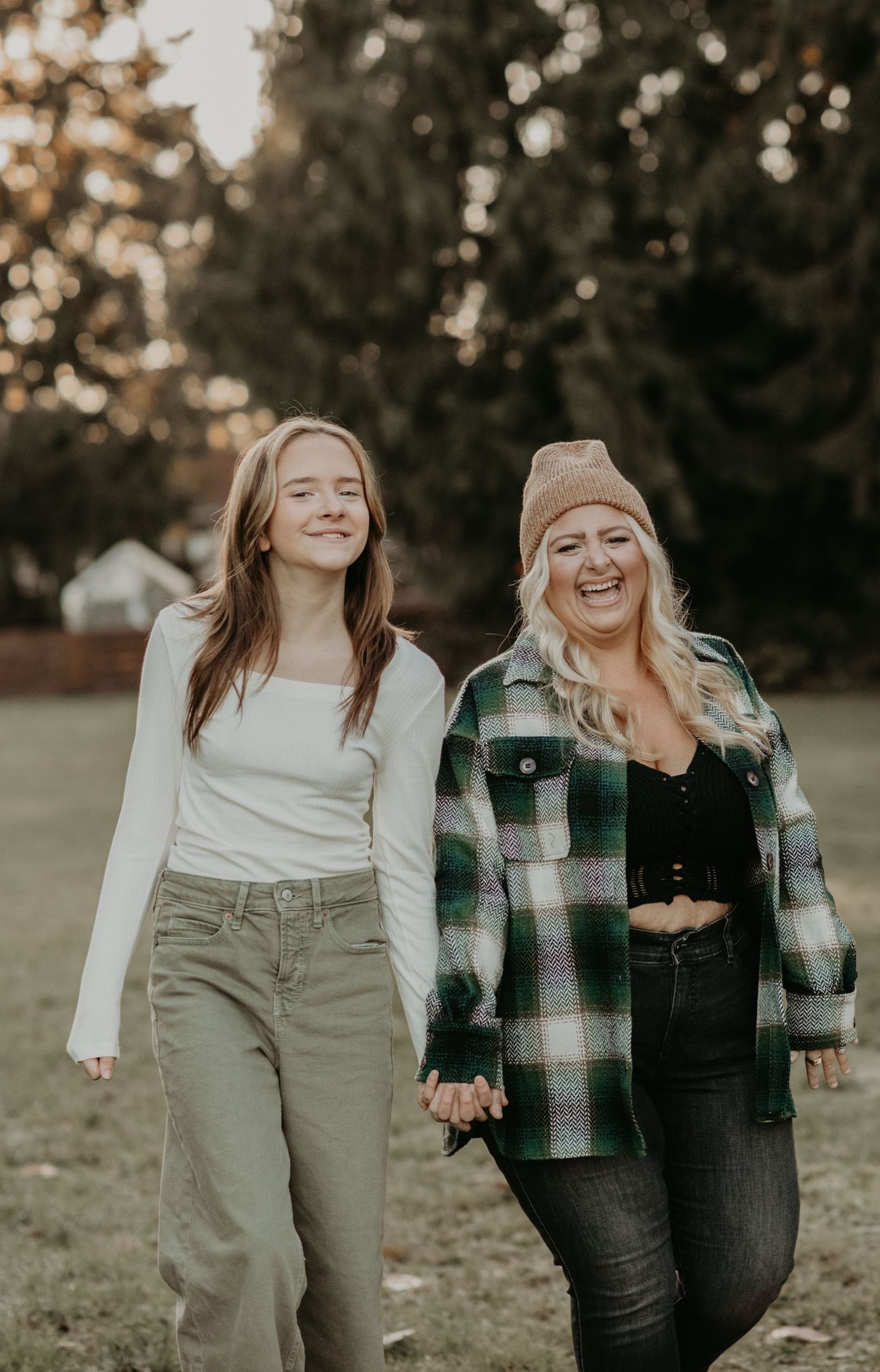 Two friends hold hands and laugh together while walking outdoors on an autumn day in casual clothing.