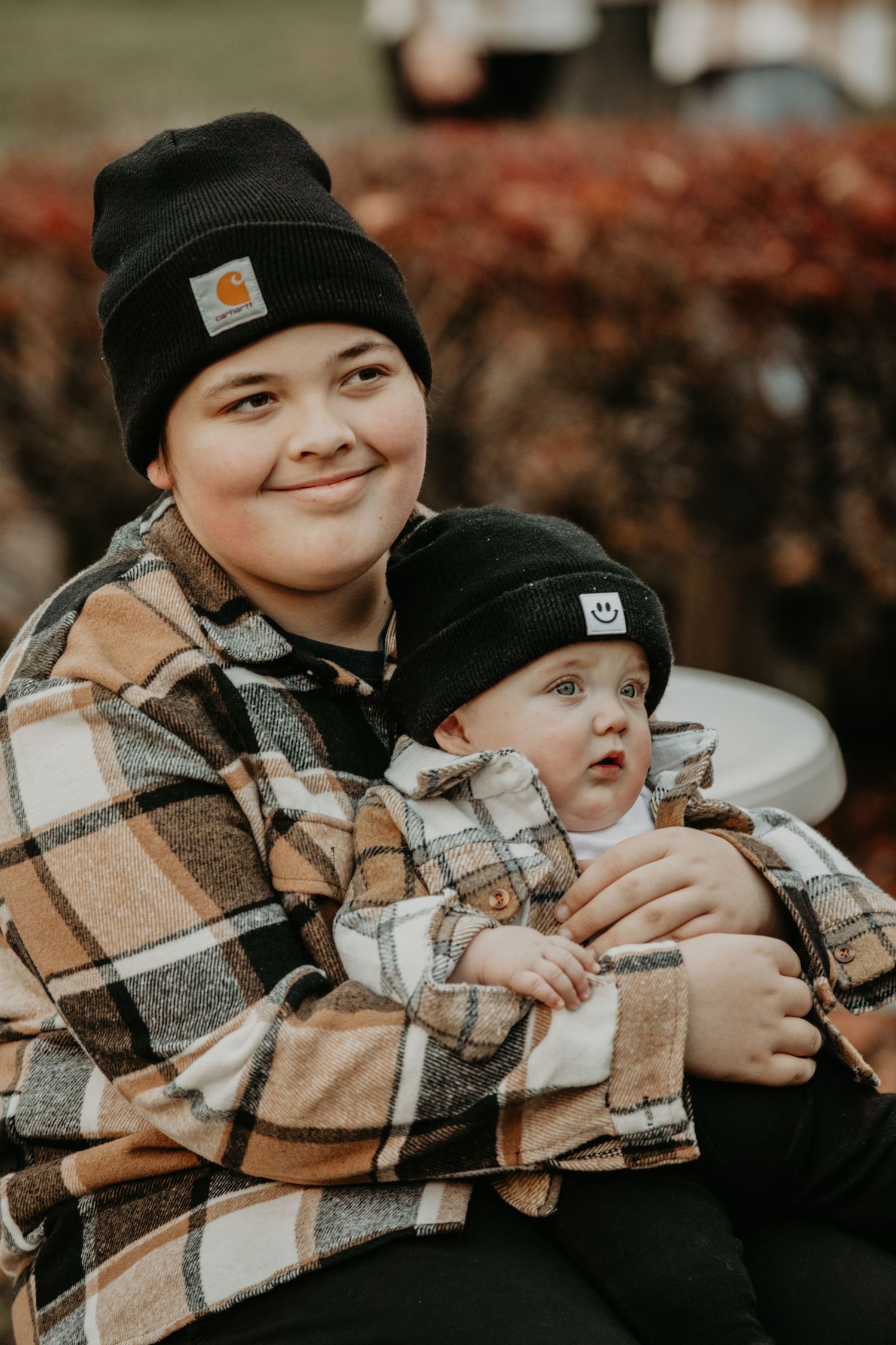 Two people wearing matching brown plaid flannel jackets and black Carhartt beanies sit close together outdoors in autumn.