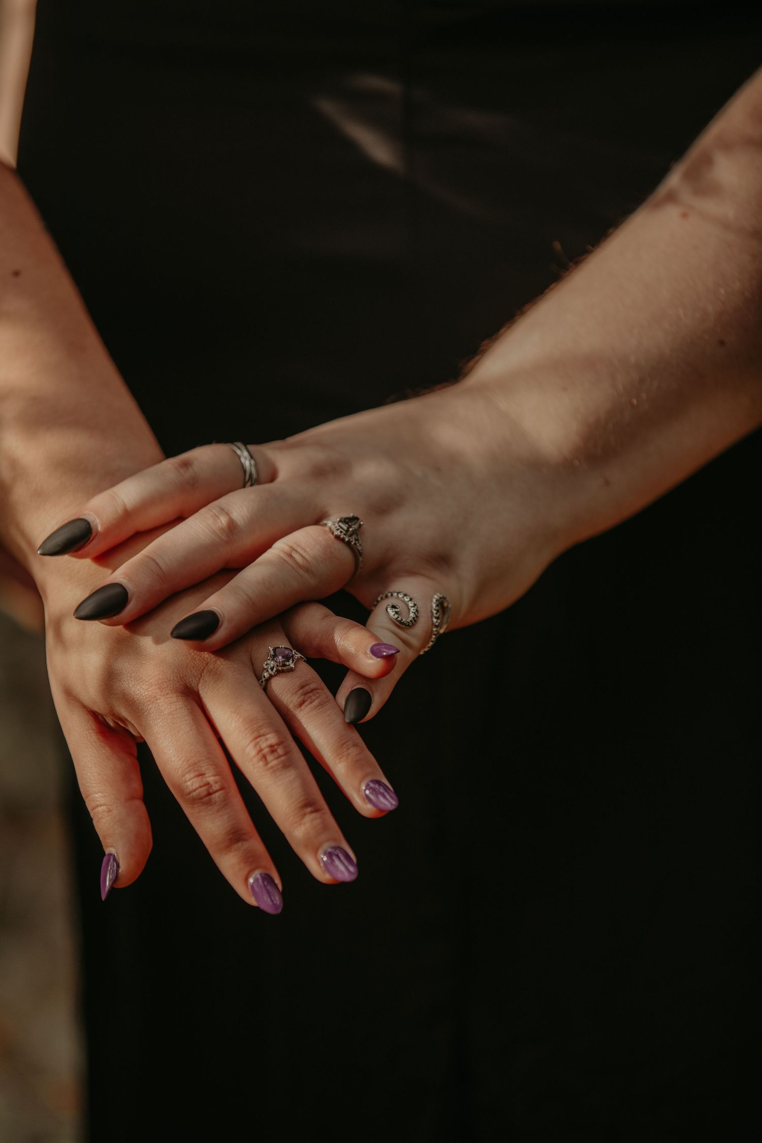 Hands adorned with rings and artistic nail designs are crossed in an intimate pose against a dark background.