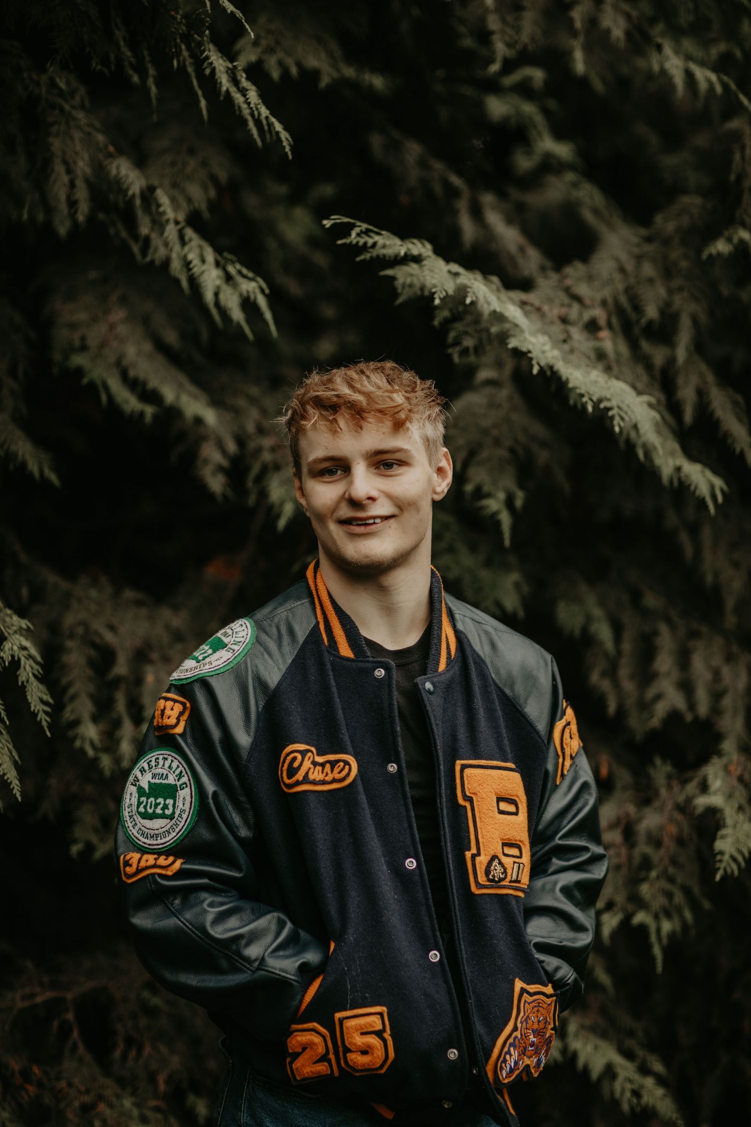 Someone in a black and orange varsity jacket with patches stands in front of evergreen branches.