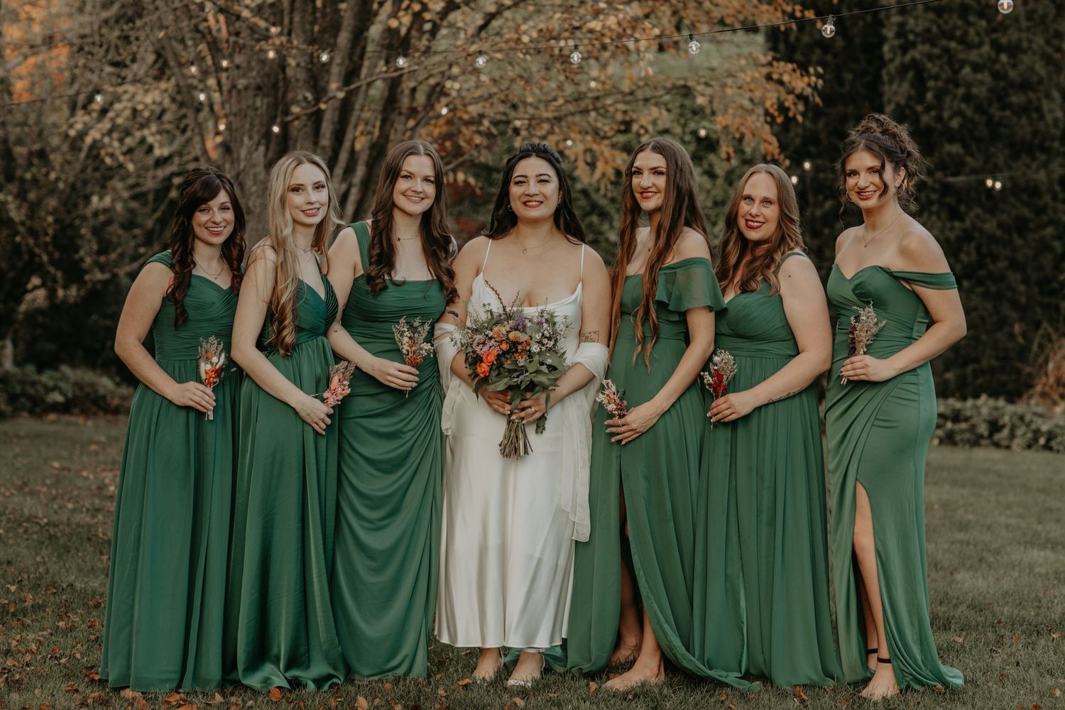 Wedding party in matching emerald green bridesmaid dresses stand together outdoors during autumn ceremony.