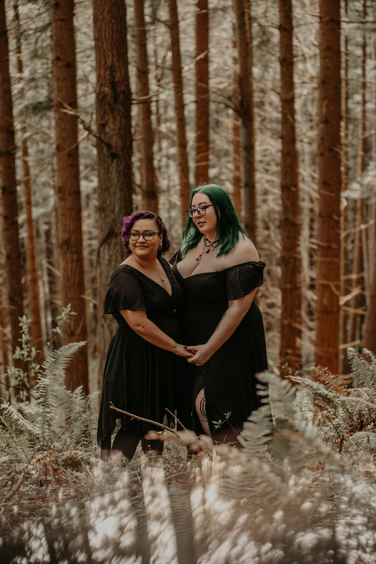 Two people in matching black dresses pose together in a moody forest setting among ferns and tall pine trees.
