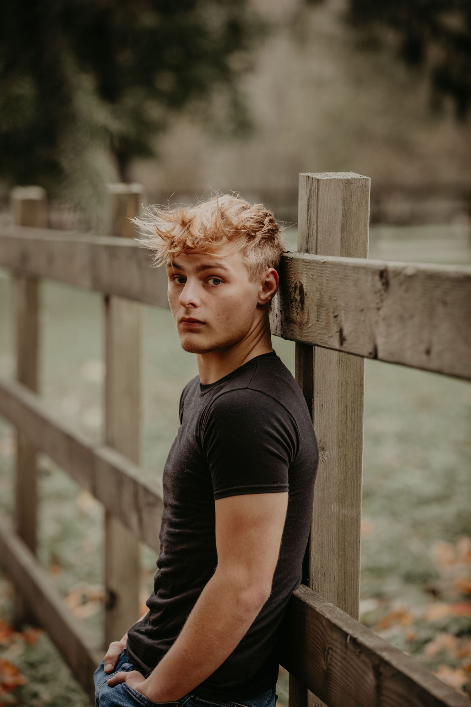 Artistic portrait of someone leaning against a wooden fence in a misty autumn setting wearing a black t-shirt.