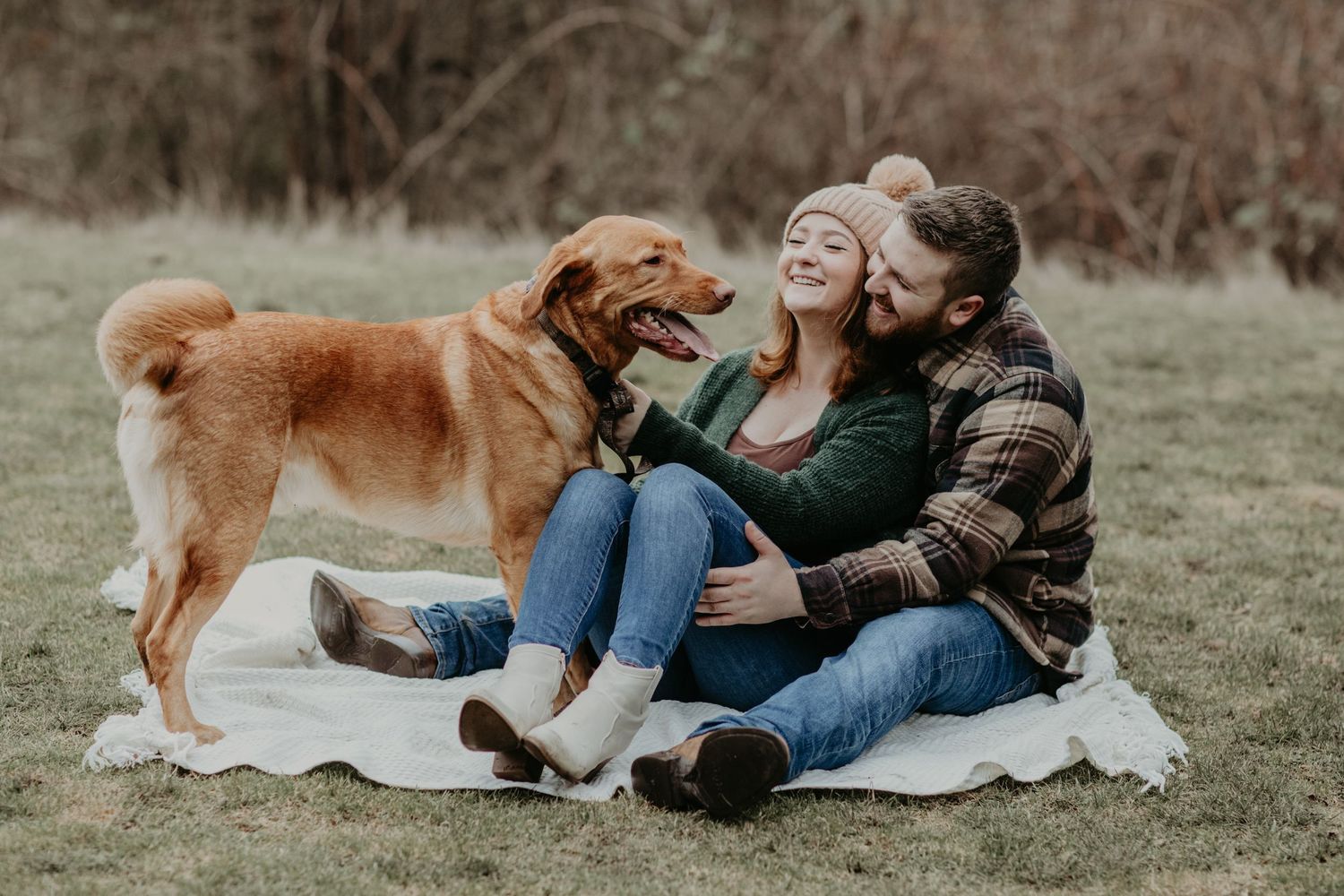 Couple sits on picnic blanket outdoors with their brown fluffy dog on a casual autumn day.