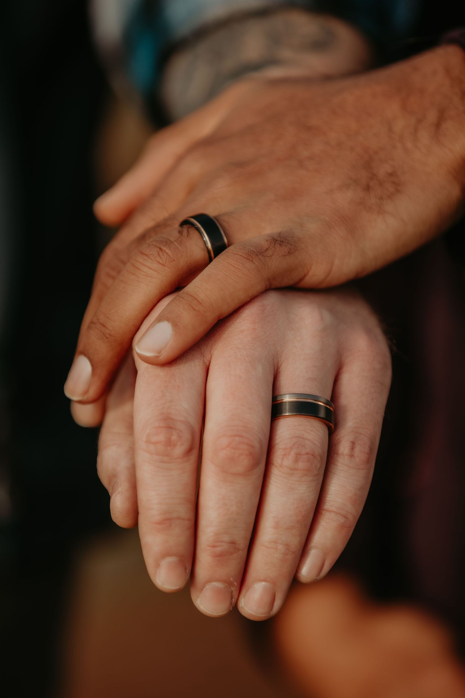 Close up of two hands with matching black wedding bands gently touching in warm lighting.