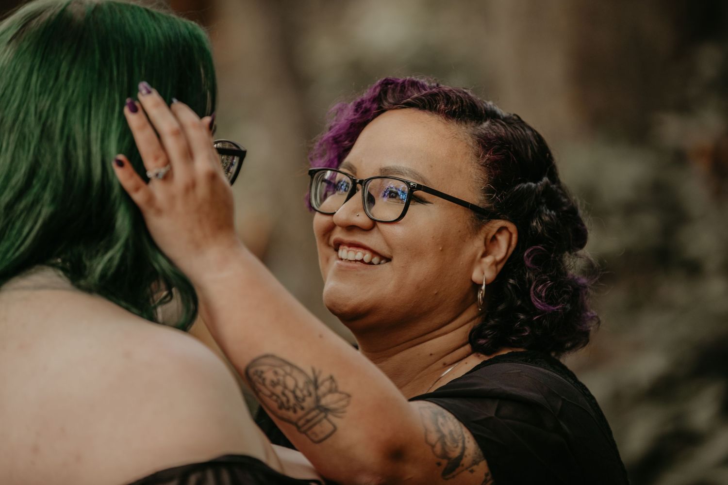 A person with glasses and curly purple-tinted hair beams with joy while wearing a black top outdoors.