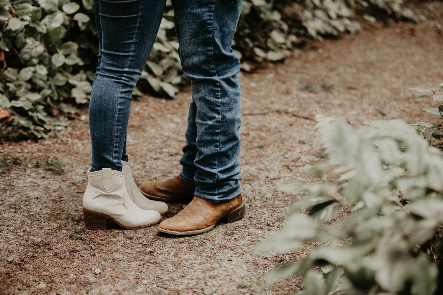 Two people in blue jeans and contrasting boots stand close together on a dirt path surrounded by foliage.
