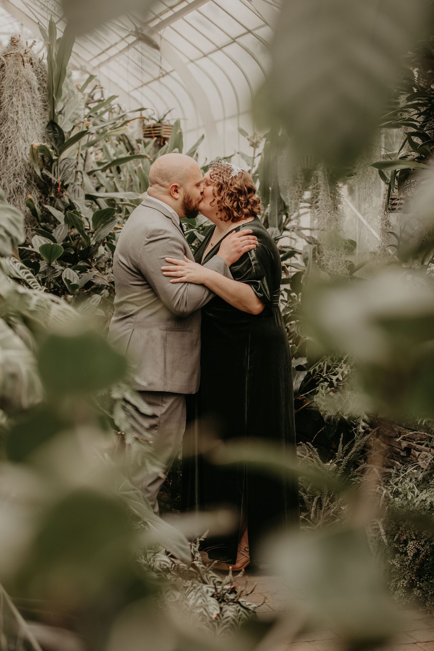A romantic couple embraces in a beautiful greenhouse setting surrounded by lush greenery and filtered natural light.