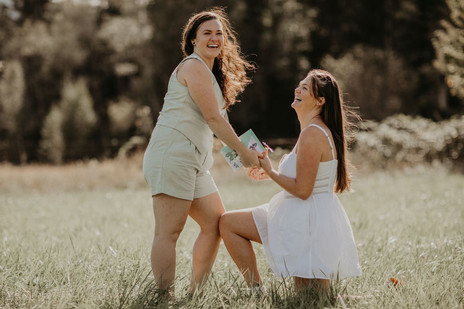 Two people share a joyful moment outdoors in a sunlit field, one wearing a white dress and one in light beige attire.