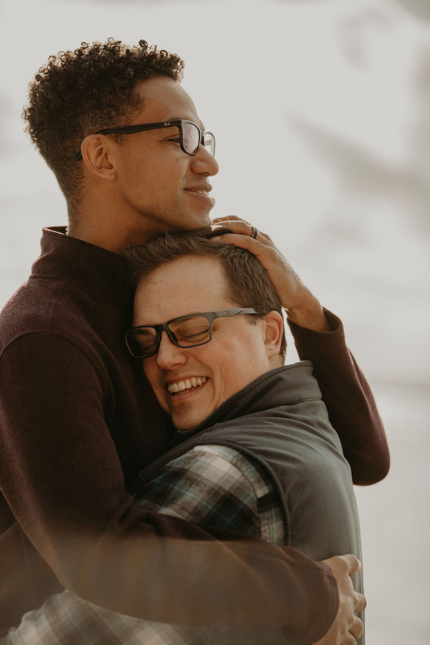 A loving couple wearing glasses share a warm embrace while smiling during a sunset photoshoot.