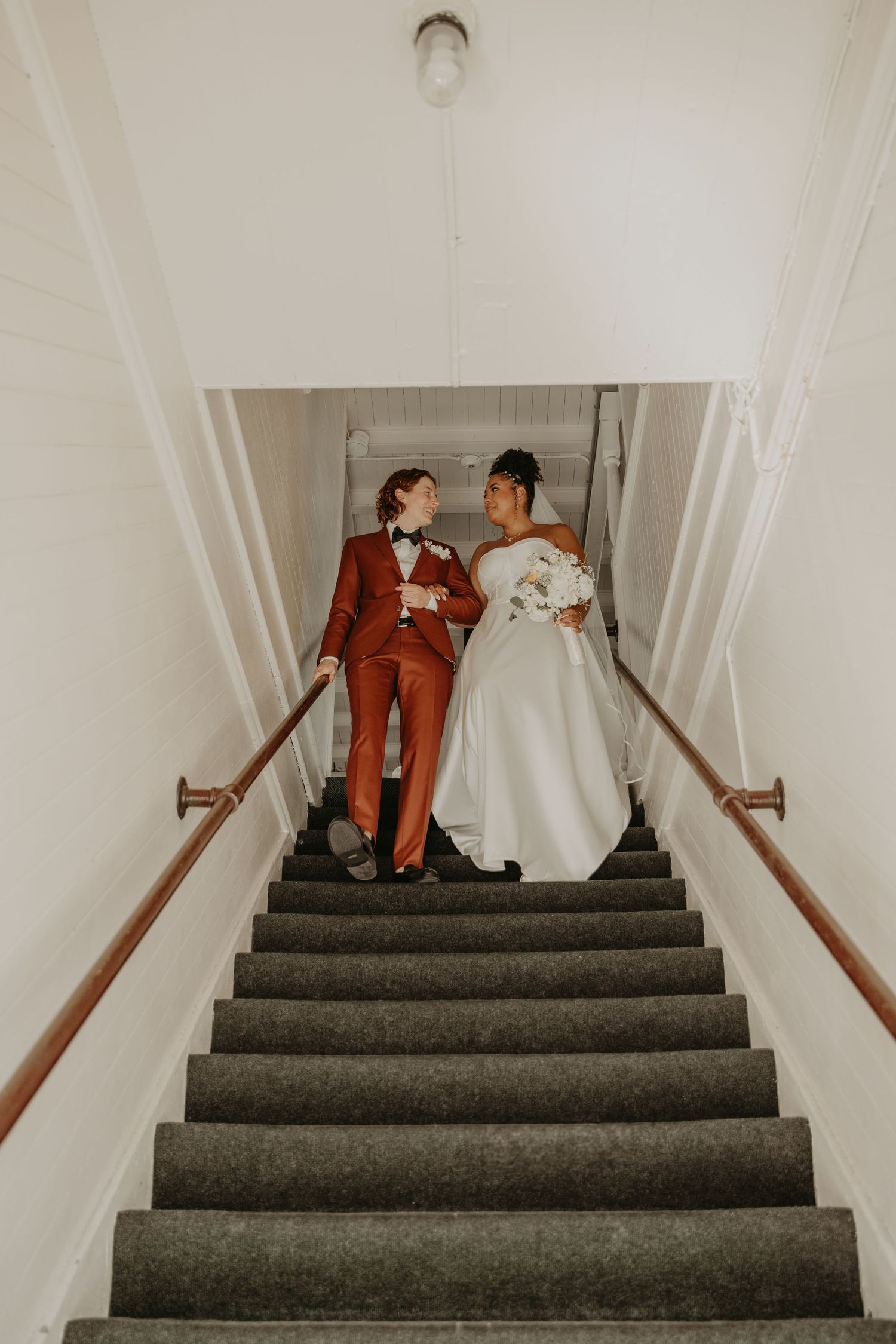 A couple in wedding attire poses together on a staircase, with one wearing a bright red suit and one in a white gown.