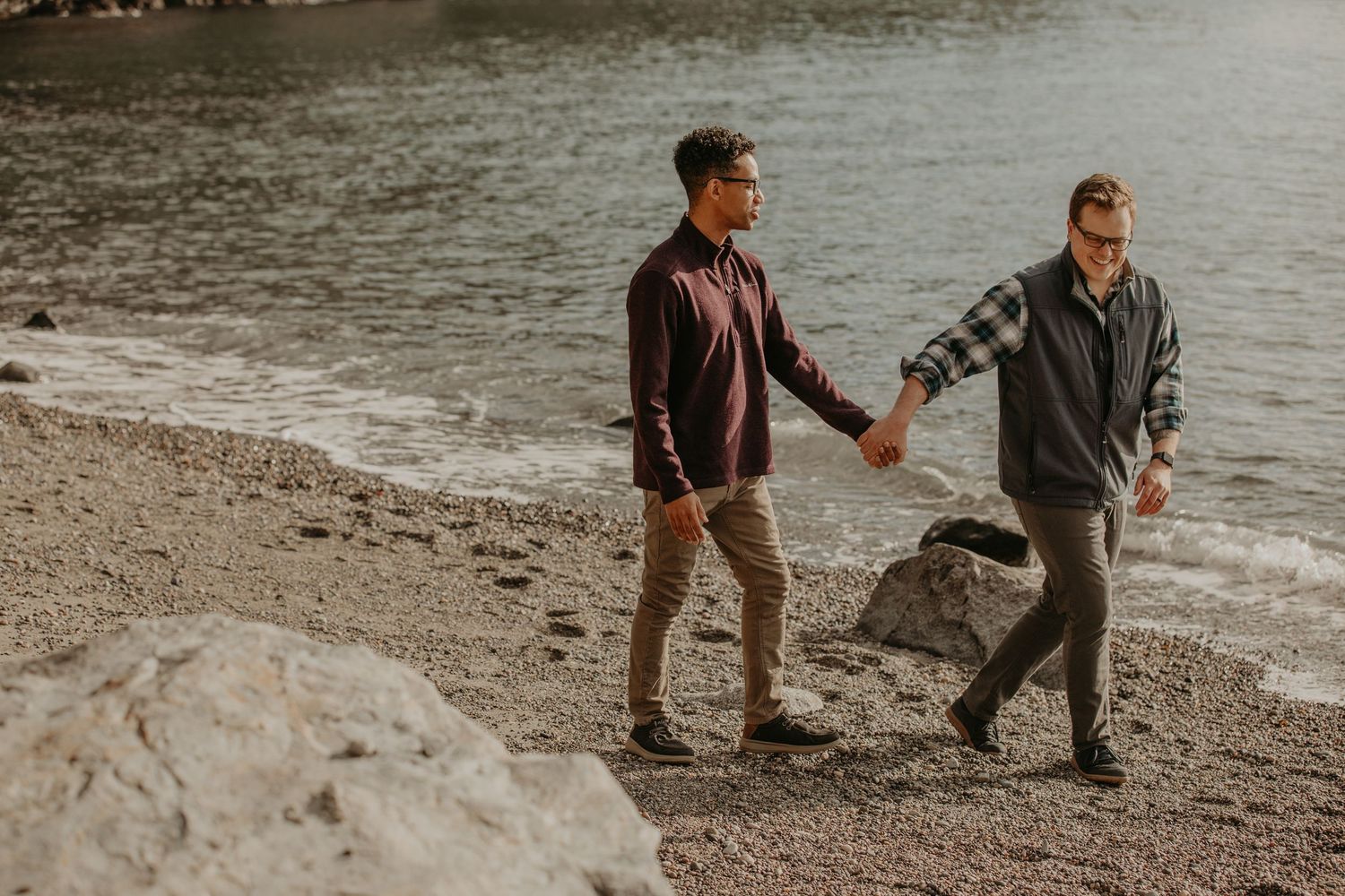 Two individuals walking hand in hand along a rocky beach shoreline during golden hour.