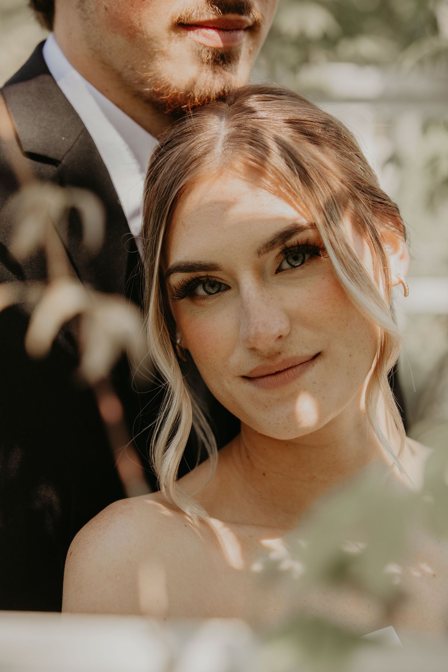 A sun-dappled wedding portrait captures a couple in an intimate outdoor setting with soft, natural lighting.