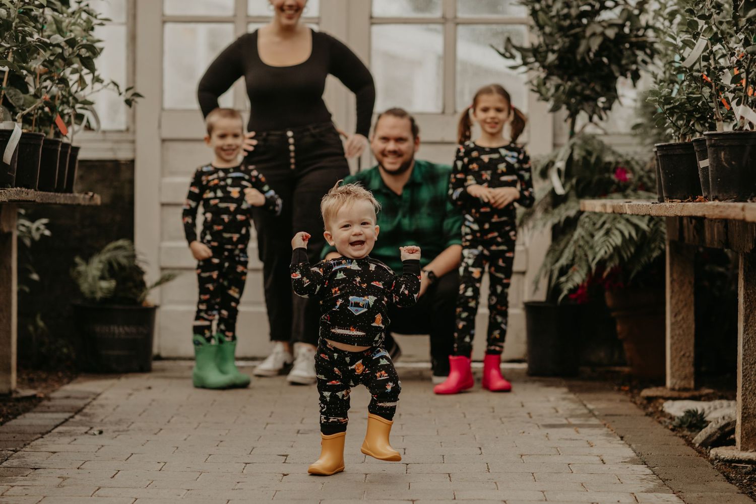 A family wearing matching patterned pajamas poses together in a greenhouse with plants and winter boots.