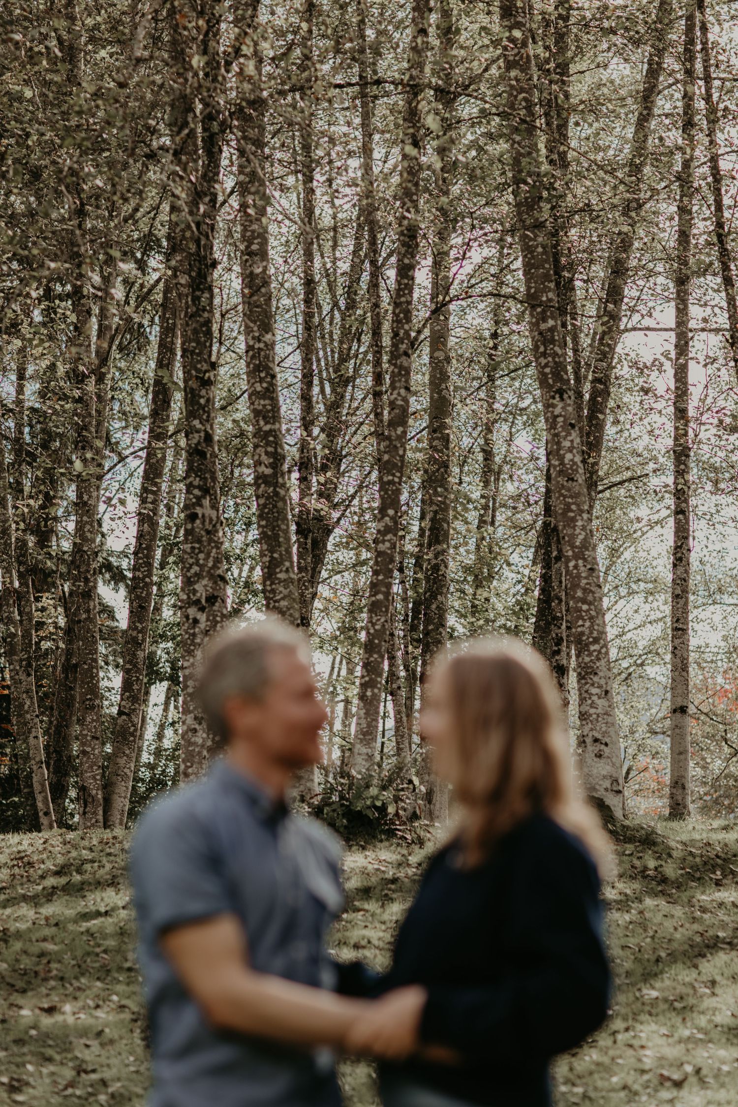 A couple shares a tender moment while standing among tall birch trees in a moody forest setting.