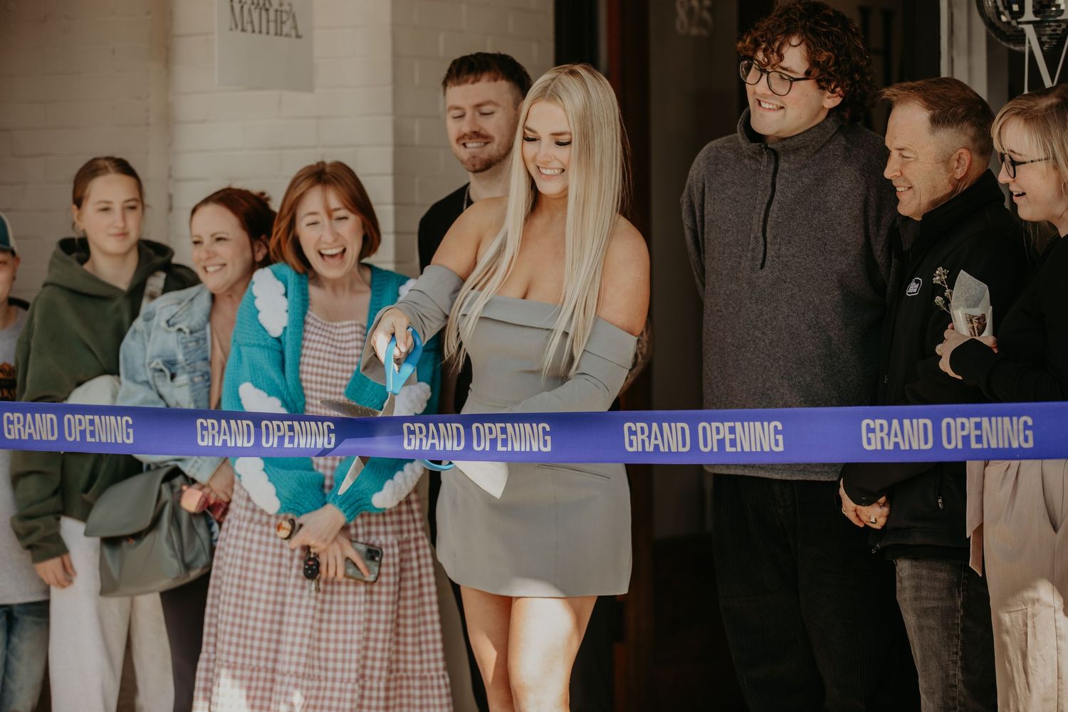 A group of people attending a grand opening ceremony gather around a blue ribbon for the cutting celebration.